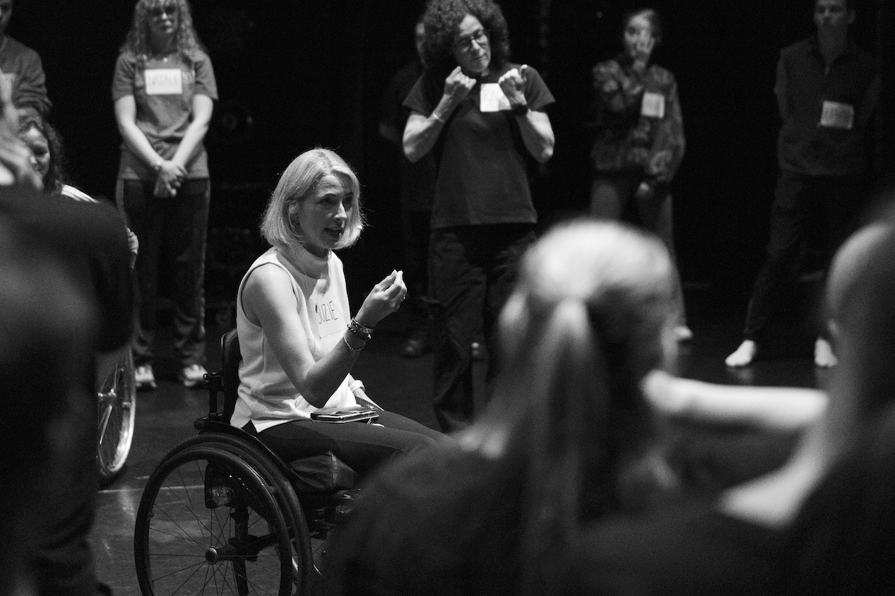 Suzie talking to dancers, giving instruction. Dancers stand behind her watching her.