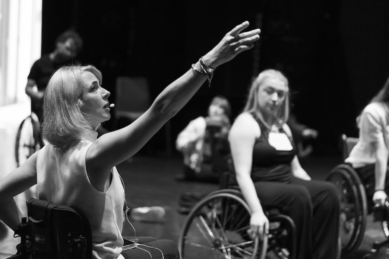 Suzie teaching a class, arm outstretched. She's looking to her finger. A female wheelchair dancer is watching her.