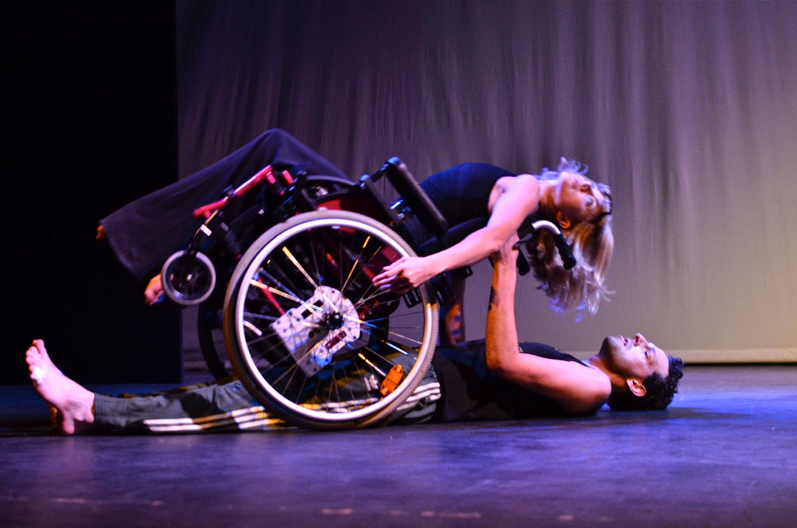 Suzie in her wheelchair tilted backwards, a male dancer lying under her on the floor, taking hold of her shoulders.