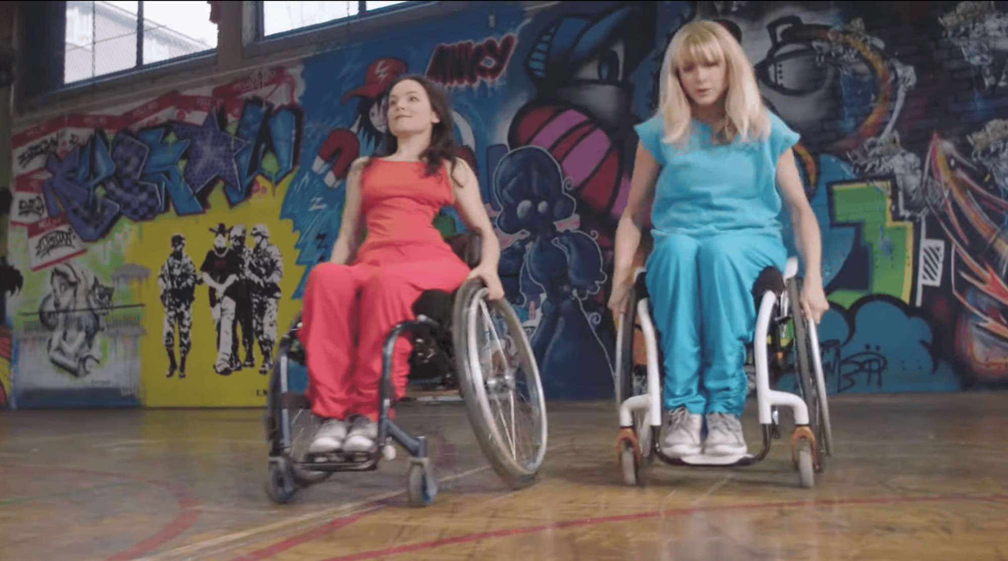 Image caption: Two women are dancing in wheelchairs in front of a brightly painted graffiti wall. Sunlight streams through high windows, casting patches of light across the wall. The overall scene feels vibrant, dynamic, and expressive, blending dance, street culture, and art. One dancer wears a red outfit and has long brown hair - Laura. The other - Suzie - wears turquoise and has long blonde hair. Both are white. Both move with energy and focus.
