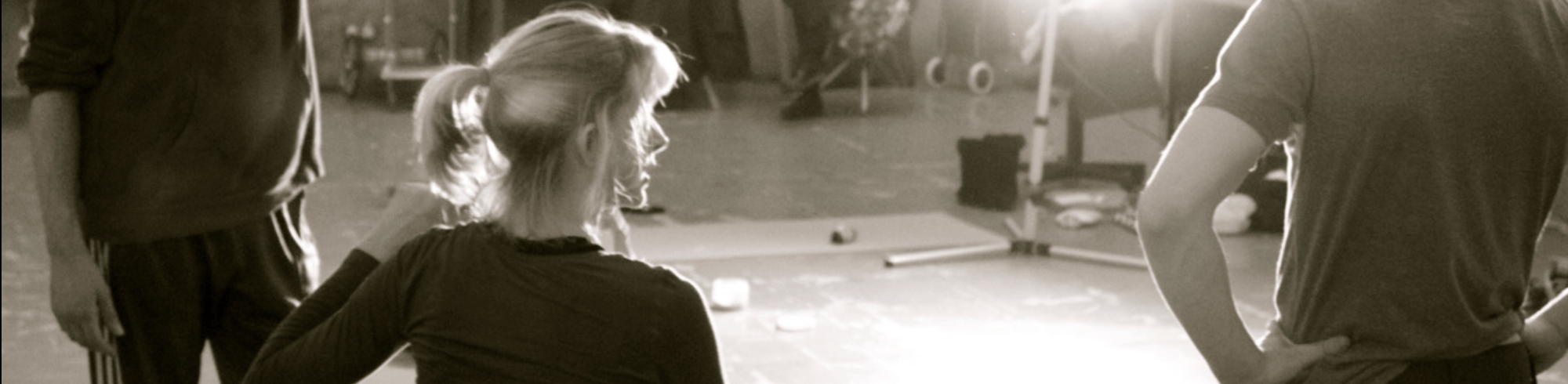 Black and white photo of Suzie teaching two male dancers in a studio. We see Suzie from behind, sitting in her wheelchair. Her face is turned to the right and her face is shown in profile. She's talking to the dancers.