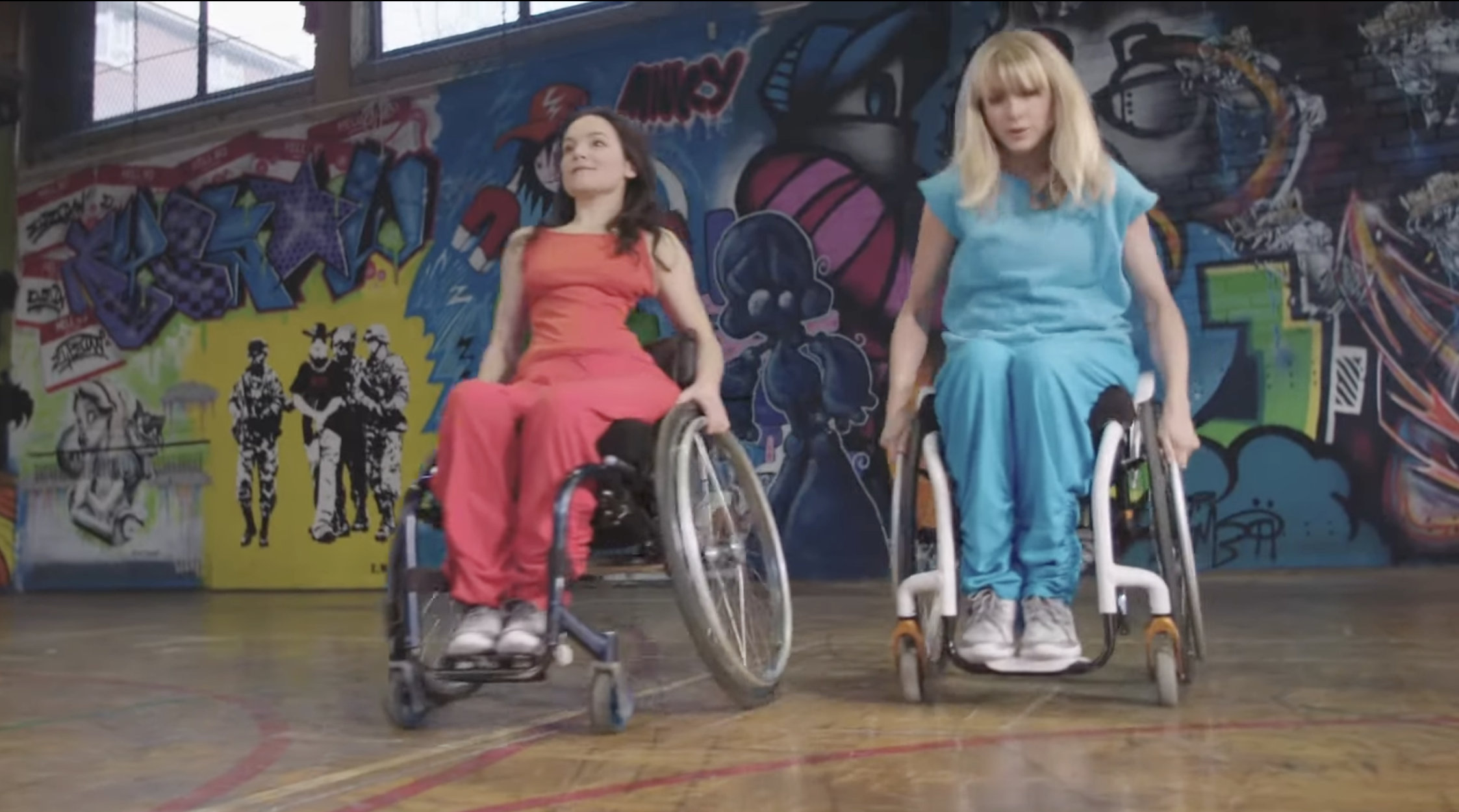 Two women are dancing in wheelchairs in front of a brightly painted graffiti wall. Sunlight streams through high windows, casting patches of light across the wall. The overall scene feels vibrant, dynamic, and expressive, blending dance, street culture, and art. One dancer wears a red outfit and has long brown hair - Laura. The other - Suzie - wears turquoise and has long blonde hair. 