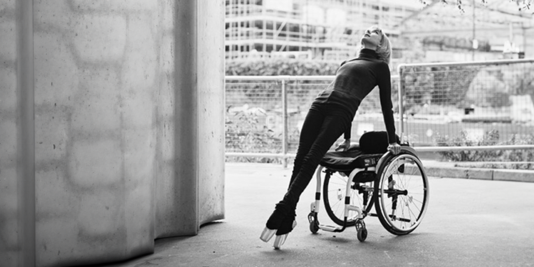 A black and white image of Suzie and her wheelchair. She is in an urban landscape - a large concrete wall is to her left and the ground is flat, grey concrete. Behind her is metal fencing, a brick wall, and a construction site beyond. Suzie has her shoulder-length hair down and wears all black with shiny ballet slippers. Her body is strong and straight, making a 45 degree angle as she presses down onto her wheel rims with straight, strong arms. She looks up to the sky and her ankles are crossed as she points her toes. She looks strong, confident and in control.