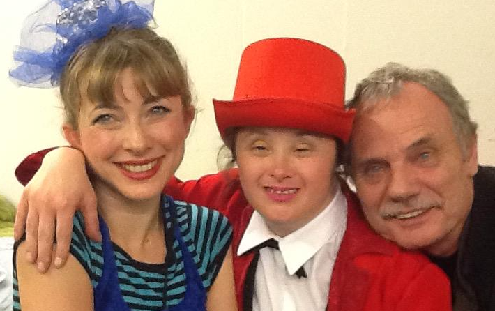 Suzie with Rosie Leak and Wolfgang. Suzie wears red lipstick and a blue velvet short dress featuring white dots and a stripy tshirt. Her arm is in a sling made from blue velvet. She is smiling. Rosie has a red top hat and tails, white shirt and black bow tie. Her arm is around Suzie and she smiles. Wolfgang rests his head on Rosie's shoulder and smiles. He has kind eyes and grey hair and moustache.